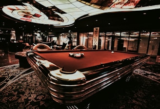 A luxurious interior features a billiard table with balls arranged on it. The ceiling has an intricate stained glass design, and the space is dimly lit, creating a sophisticated atmosphere. Comfortable chairs are placed around the area, with some decorative plants and a lamp adding to the elegance.